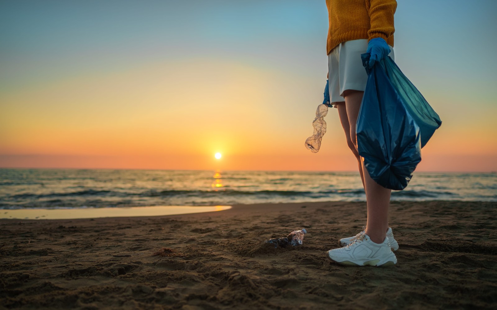 Person picking up beach litter at sunset, holding a trash bag.