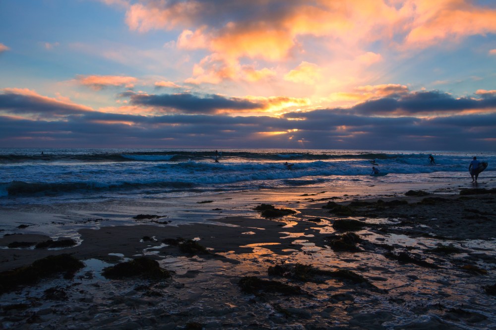 Sunset over a beach with waves and scattered clouds, silhouetted people in the distance.