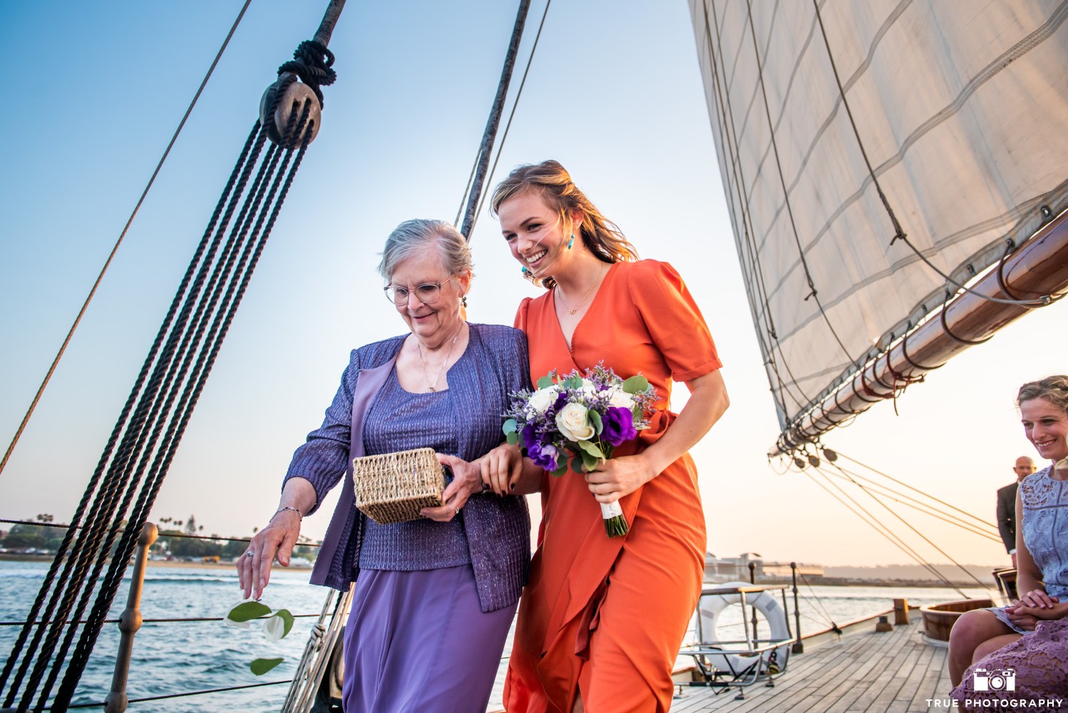 Two women on a sailboat deck, one in orange holding flowers, the other in purple holding a basket.