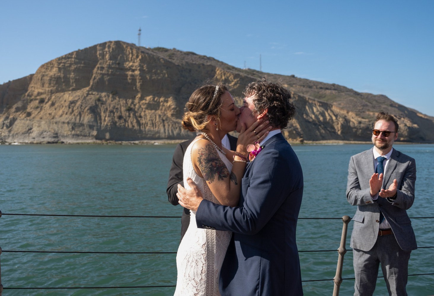 Couple kissing at outdoor wedding by water, with man clapping, and rocky hill in background.