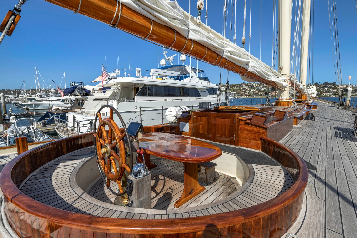 Wooden sailboat deck with steering wheel, moored by modern yachts in a marina.