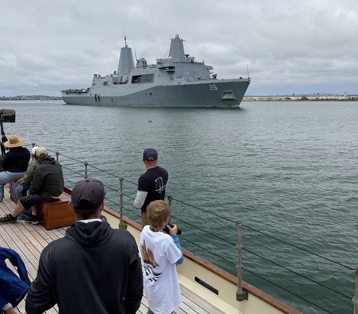 Large gray naval ship on water, viewed from a deck with onlookers.