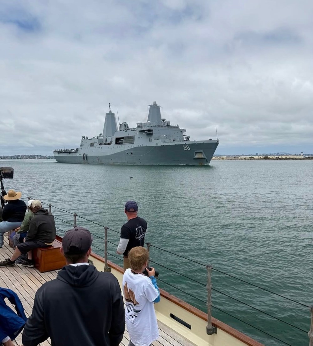 Large gray naval ship on water, viewed from a deck with onlookers.