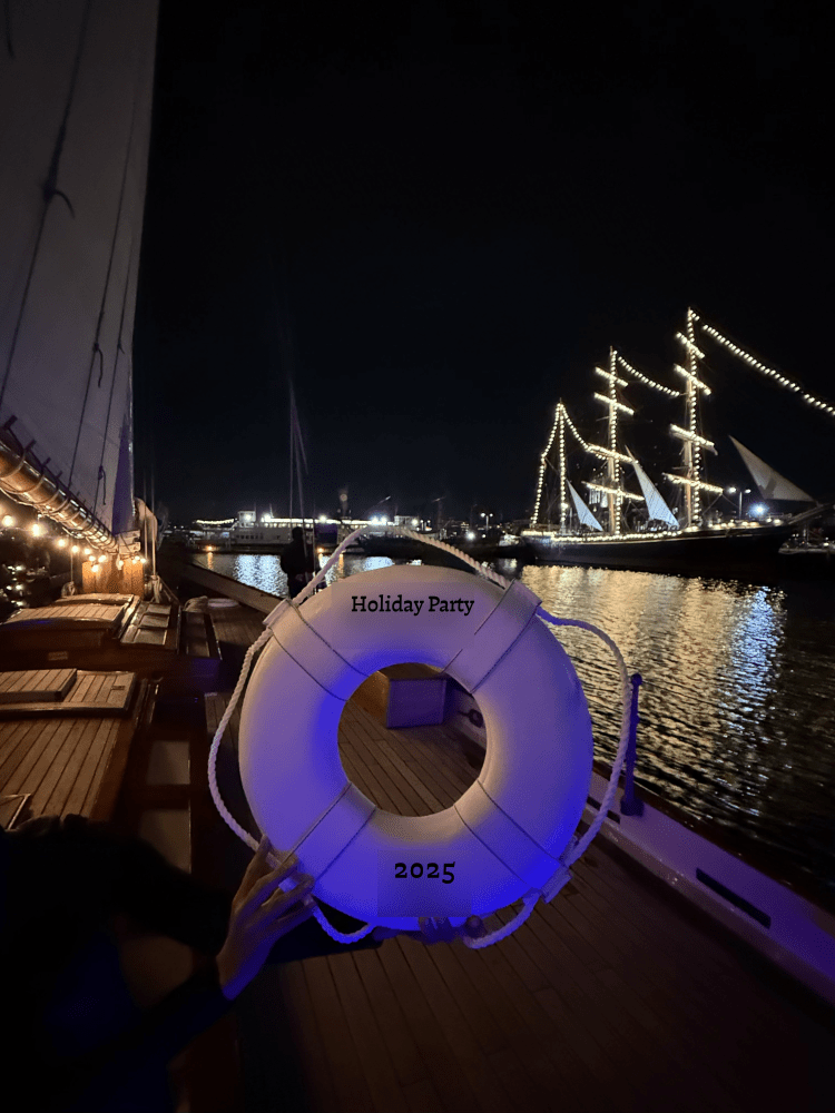 Nighttime boat party with a lit lifebuoy labeled 'Holiday Party 2025' held up on a deck near illuminated ships.