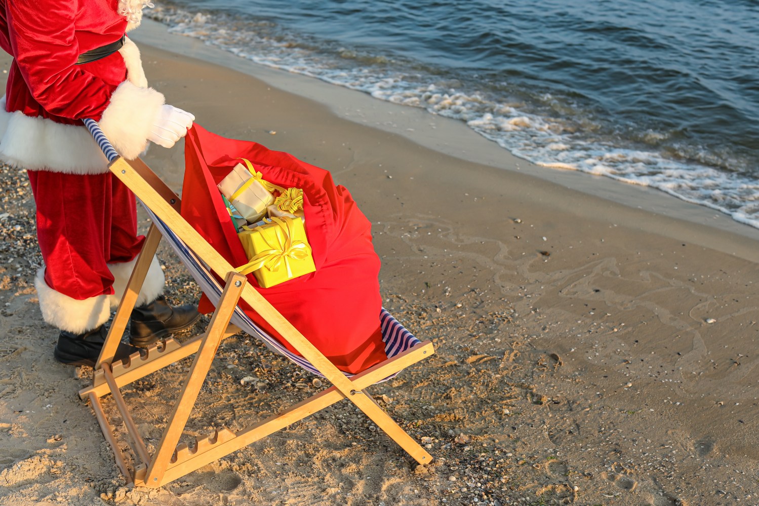 Santa Claus with gifts in a chair on a beach by the ocean.