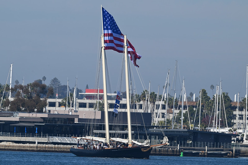 Sailing ship with American flag in harbor, surrounded by smaller boats and buildings.