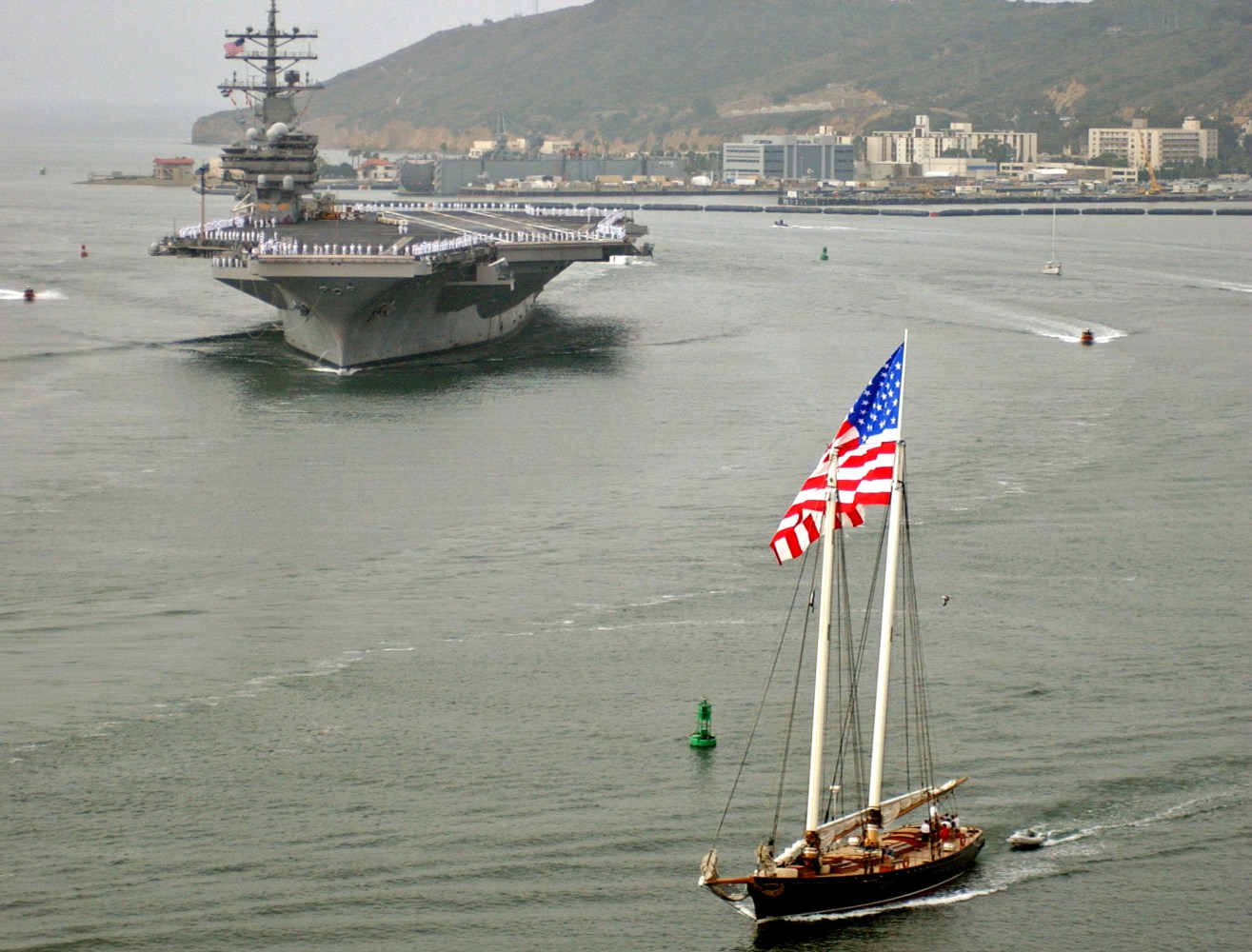 Aircraft carrier and sailboat with US flag in a harbor.
