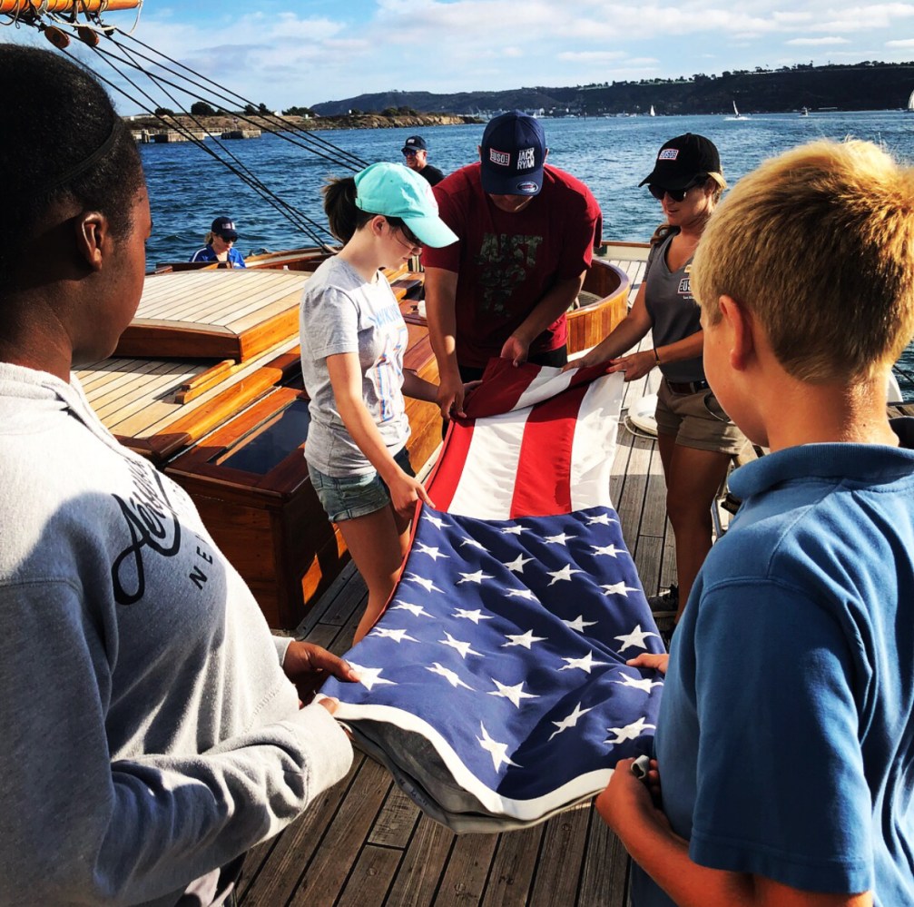 People folding a U.S. flag on a wooden boat deck, near water.