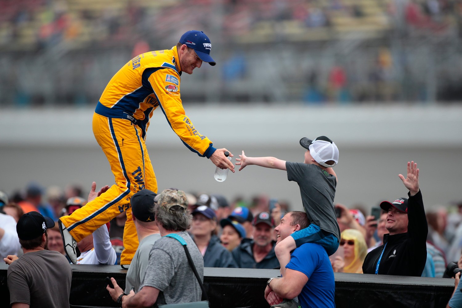 Racecar driver in yellow suit hands item to young fan in crowd.